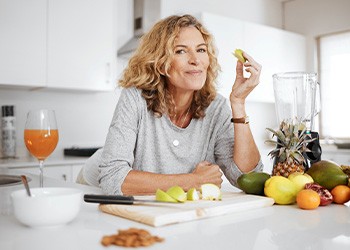 Woman leaning on counter with various fruits eating a sliced apple